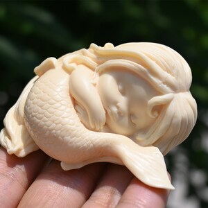 May include: A small, white, sleeping mermaid figurine with a detailed tail and long hair. The figurine is made of a smooth, ivory-like material.