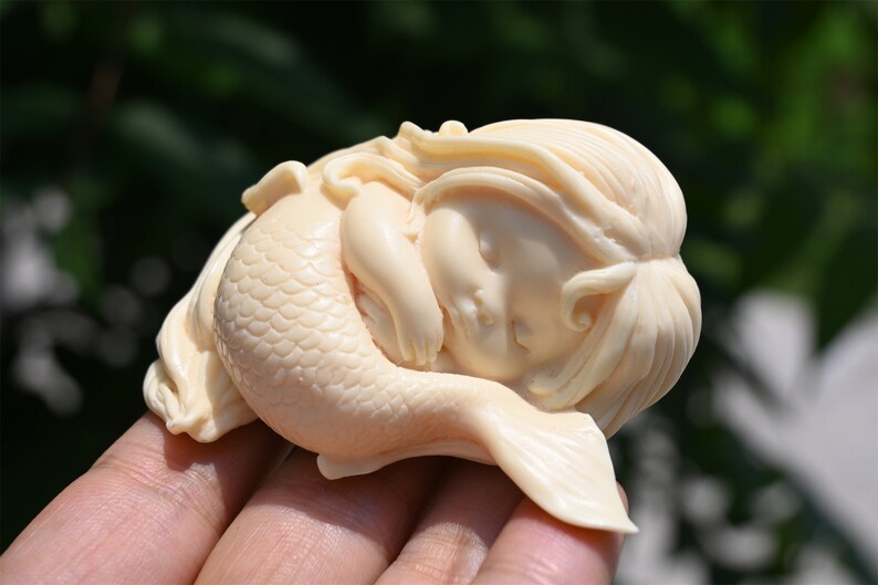 May include: A small, white, sleeping mermaid figurine with a detailed tail and long hair. The figurine is made of a smooth, ivory-like material.