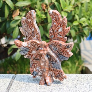 May include: A carved stone sculpture of a bird with outstretched wings. The sculpture is primarily reddish-brown with gray and white accents. The wings and body feature detailed carvings, and the sculpture is set against a blurred green background.