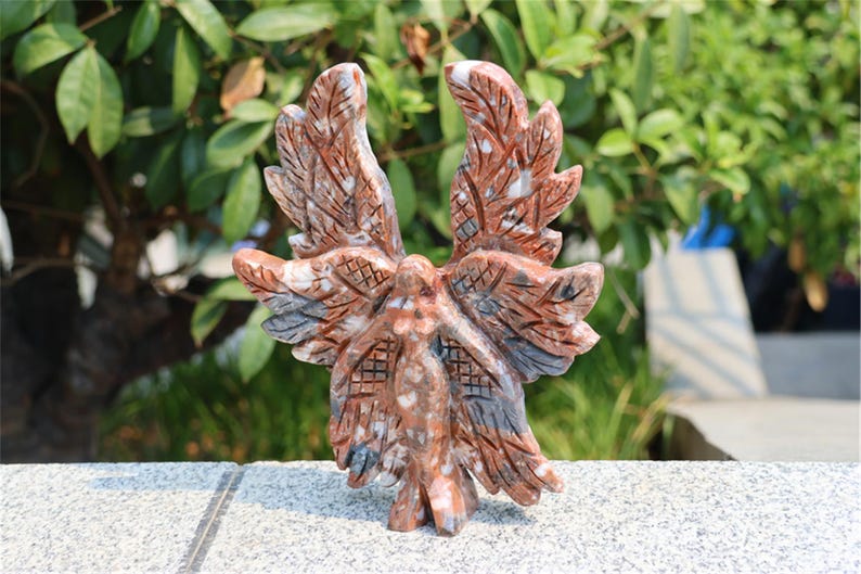 May include: A carved stone sculpture of a bird with outstretched wings. The sculpture is primarily reddish-brown with gray and white accents. The wings and body feature detailed carvings, and the sculpture is set against a blurred green background.