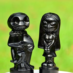 May include: Two black figurines of stylized characters, one male and one female, stand side-by-side against a blurred green backdrop. The male figure has a round head with large eye sockets and a cloak. The female figure has long hair and a skeletal grin.