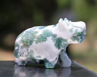 Puede incluir: Pequeña figura de hipopótamo de piedra tallada. El hipopótamo es blanco con patrones moteados verdes y verde azulado. La figura está en posición de pie con la boca ligeramente abierta. El hipopótamo se exhibe sobre una superficie oscura con un fondo borroso.