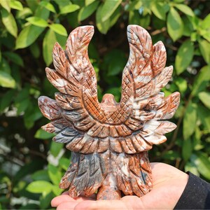 May include: A carved stone eagle sculpture with outstretched wings. The sculpture is primarily brown and gray, with detailed feather patterns. The eagle is held in a hand against a backdrop of green foliage.