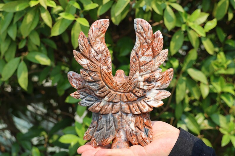 May include: A carved stone eagle sculpture with outstretched wings. The sculpture is primarily brown and gray, with detailed feather patterns. The eagle is held in a hand against a backdrop of green foliage.