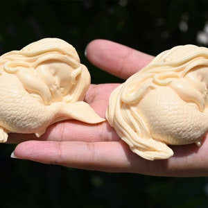 May include: Two small, cream-colored, sleeping mermaid figurines with long hair and detailed scales. The mermaids are curled up in a sleeping position.