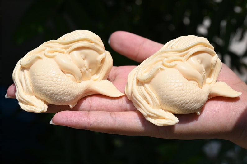 May include: Two small, cream-colored, sleeping mermaid figurines with long hair and detailed scales. The mermaids are curled up in a sleeping position.