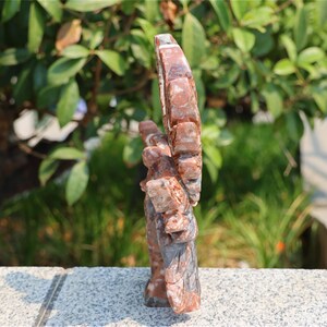 May include: A carved stone sculpture of a hand with an open palm and raised fingers. The sculpture is primarily reddish-brown with gray and white accents. The background is blurred, showing green foliage.