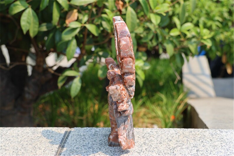 May include: A carved stone sculpture of a hand with an open palm and raised fingers. The sculpture is primarily reddish-brown with gray and white accents. The background is blurred, showing green foliage.