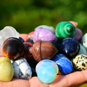 May include: A collection of polished gemstone acorns in various colours, including clear, pink, green, brown, purple, blue, and white. The acorns are displayed in a hand.