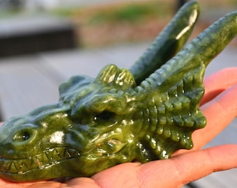 Op de afbeelding: Een groene jade drakenhoofdsculptuur. De draak heeft een gedetailleerd gezicht met scherpe tanden en twee hoorns. De sculptuur wordt in een hand gehouden, wat de ingewikkelde gravure en de gepolijste afwerking benadrukt.