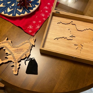 May include: A wooden game board shaped like the outline of a lake, with the words "Lindy Lake, Chequamegon Lake, WI, Established 2014" engraved on the board. The board is in a wooden box with a glass top.