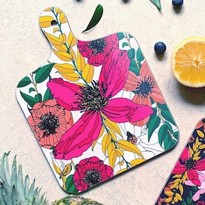 May include: Two floral patterned cutting boards. The first board has a white background with pink, yellow, and green floral designs. The second board has a black background with pink, white, and green floral designs. Both boards have a handle.