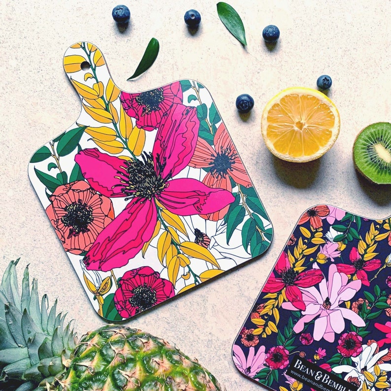 May include: Two floral patterned cutting boards. The first board has a white background with pink, yellow, and green floral designs. The second board has a black background with pink, white, and green floral designs. Both boards have a handle.