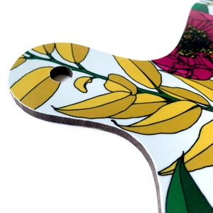 May include: A white cutting board with a floral design featuring yellow leaves and pink flowers. The board has a hole in the top for hanging.