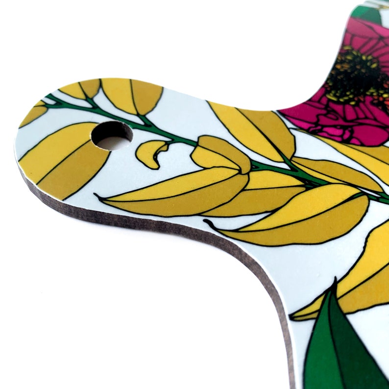 May include: A white cutting board with a floral design featuring yellow leaves and pink flowers. The board has a hole in the top for hanging.