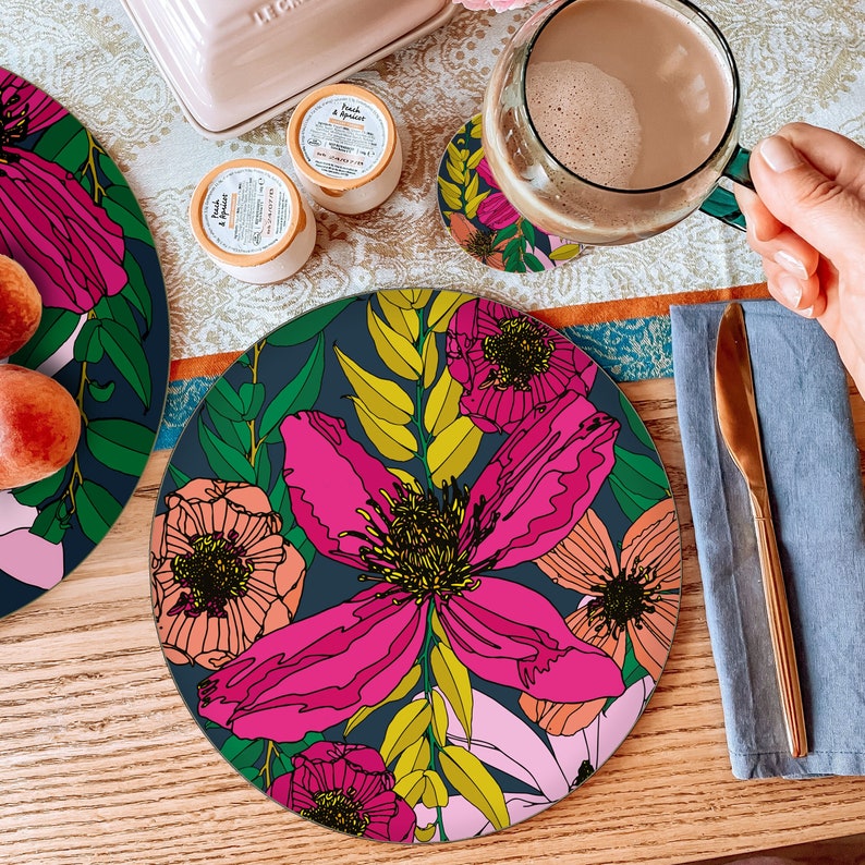 May include: Round floral print placemat with pink, orange, and yellow flowers on a dark blue background. The placemat is made of cork and is perfect for adding a touch of style to your dining table.