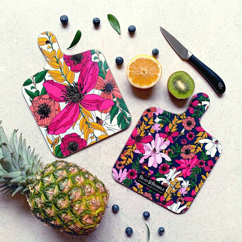 May include: Two floral patterned cutting boards, one with pink and white flowers on a white background and the other with pink, red, and white flowers on a black background. The boards are made of wood and have a beveled edge. The text "Bean & Bramble" is printed on the black board.