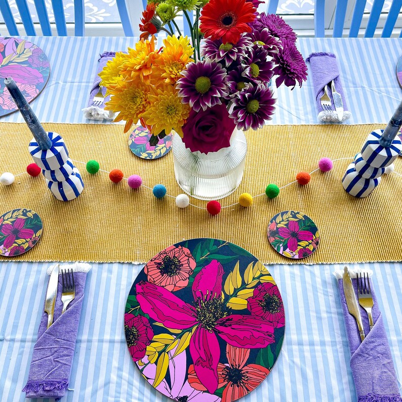 May include: A table setting with a yellow and blue striped tablecloth, a yellow placemat, and a floral print placemat. The centerpiece is a bouquet of pink, yellow, and purple flowers in a clear glass vase. There are also colorful pom poms strung across the table.
