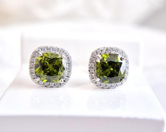 May include: A pair of square-cut, olive-green gemstone stud earrings. Each earring is surrounded by a halo of small, clear gemstones set in a silver-colored metal. The earrings are displayed on a white surface.