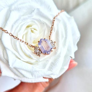 May include: A rose gold necklace featuring a rectangular, light purple gemstone pendant. The pendant is flanked by smaller, clear gemstones. The necklace is displayed on a white rose.