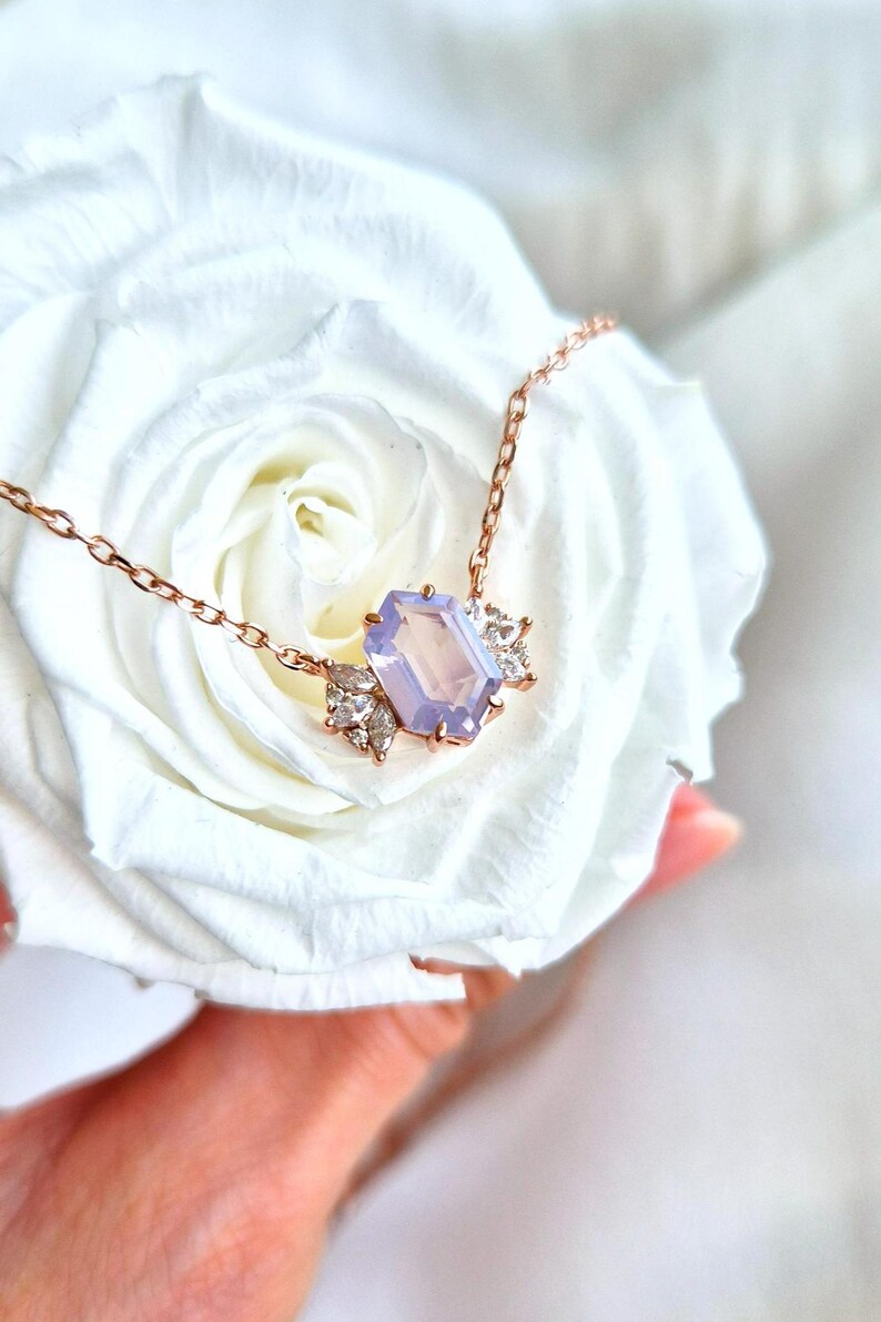May include: A rose gold necklace featuring a rectangular, light purple gemstone pendant. The pendant is flanked by smaller, clear gemstones. The necklace is displayed on a white rose.