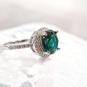 May include: A silver ring with a large, round, emerald green gemstone surrounded by a halo of smaller, clear gemstones.