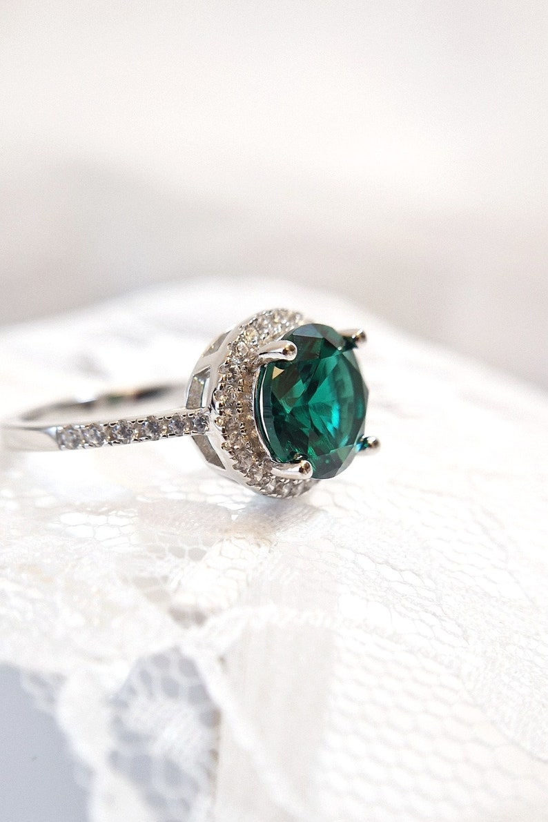 May include: A silver ring with a large, round, emerald green gemstone surrounded by a halo of smaller, clear gemstones.