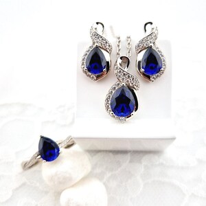 May include: A silver jewelry set with a teardrop-shaped blue gemstone pendant, matching earrings, and a ring. The set is displayed on a white background.