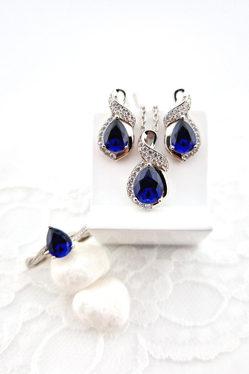 May include: A silver jewelry set with a teardrop-shaped blue gemstone pendant, matching earrings, and a ring. The set is displayed on a white background.
