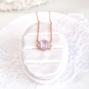 May include: A rose gold necklace with a light purple, hexagonal gemstone pendant. The pendant is flanked by two smaller, clear crystal accents. The necklace is displayed on a white, velvet jewelry box.