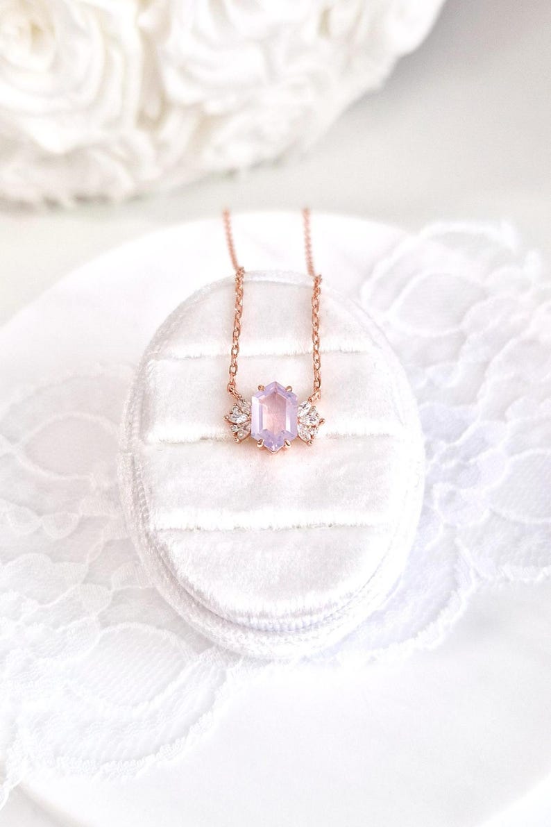 May include: A rose gold necklace with a light purple, hexagonal gemstone pendant. The pendant is flanked by two smaller, clear crystal accents. The necklace is displayed on a white, velvet jewelry box.