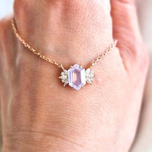 May include: A delicate gold necklace featuring a light purple, hexagonal gemstone pendant. The pendant is flanked by two clusters of small, sparkling white stones. The necklace is displayed on a person's hand.