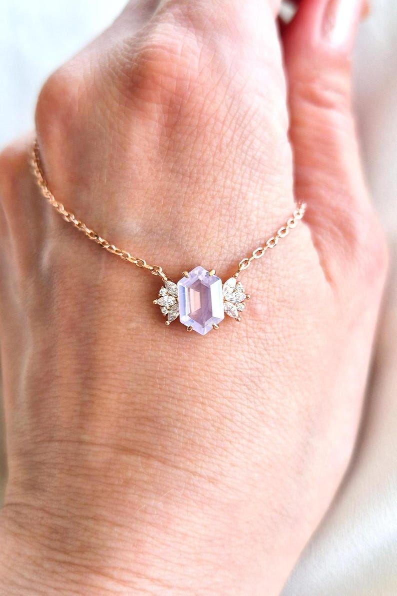 May include: A delicate gold necklace featuring a light purple, hexagonal gemstone pendant. The pendant is flanked by two clusters of small, sparkling white stones. The necklace is displayed on a person's hand.