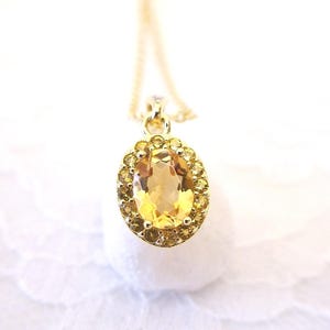 K&ouml;nnte beinhalten: Eine goldene Anh&auml;ngerkette mit einem gro&szlig;en, oval geschliffenen gelben Edelstein, der von kleineren gelben Steinen umgeben ist. Der Anh&auml;nger h&auml;ngt an einer zarten Goldkette, vor einem weichen, wei&szlig;en Hintergrund. Der Schmuck ist ein klassisches und elegantes Accessoire.