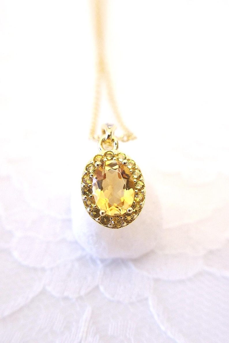 K&ouml;nnte beinhalten: Eine goldene Anh&auml;ngerkette mit einem gro&szlig;en, oval geschliffenen gelben Edelstein, der von kleineren gelben Steinen umgeben ist. Der Anh&auml;nger h&auml;ngt an einer zarten Goldkette, vor einem weichen, wei&szlig;en Hintergrund. Der Schmuck ist ein klassisches und elegantes Accessoire.
