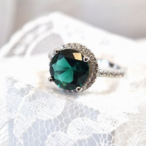 May include: A silver ring with a large, round, emerald green gemstone surrounded by a halo of smaller, clear gemstones.