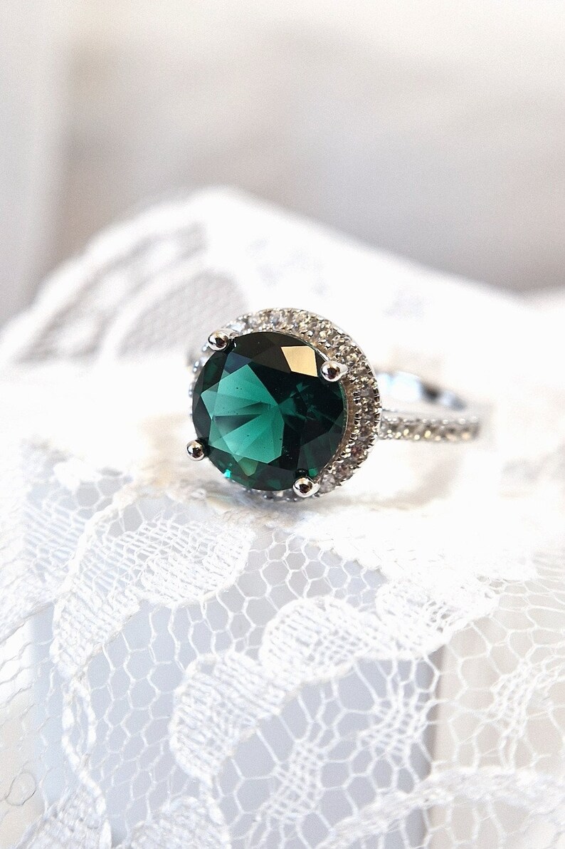 May include: A silver ring with a large, round, emerald green gemstone surrounded by a halo of smaller, clear gemstones.