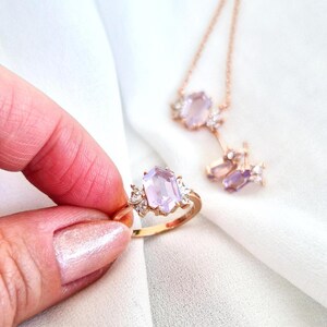 Peut inclure: Une bague en or avec une grande pierre rectangulaire violet clair, flanqu&eacute;e de petites pierres transparentes. Un collier et des boucles d'oreilles assortis avec des pierres pr&eacute;cieuses similaires sont &eacute;galement visibles, sur fond blanc.