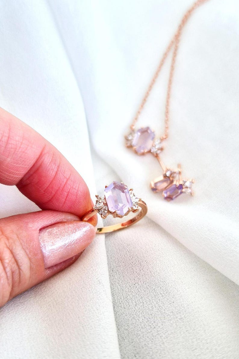Peut inclure: Une bague en or avec une grande pierre rectangulaire violet clair, flanqu&eacute;e de petites pierres transparentes. Un collier et des boucles d'oreilles assortis avec des pierres pr&eacute;cieuses similaires sont &eacute;galement visibles, sur fond blanc.