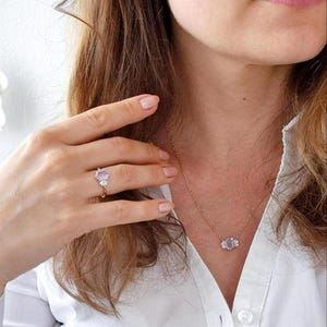 Peut inclure: Ensemble de bijoux en or comprenant un collier, une bague et des boucles d'oreilles, orn&eacute;s de pierres pr&eacute;cieuses violet clair. La bague est port&eacute;e &agrave; la main, le collier et les boucles d'oreilles sont port&eacute;s. L'ensemble est port&eacute; avec une chemise blanche.