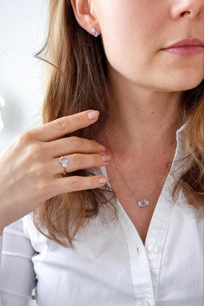 Peut inclure: Ensemble de bijoux en or comprenant un collier, une bague et des boucles d'oreilles, orn&eacute;s de pierres pr&eacute;cieuses violet clair. La bague est port&eacute;e &agrave; la main, le collier et les boucles d'oreilles sont port&eacute;s. L'ensemble est port&eacute; avec une chemise blanche.
