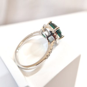 May include: A silver ring with a large, emerald green gemstone surrounded by smaller, clear gemstones. The ring is set on a white background.