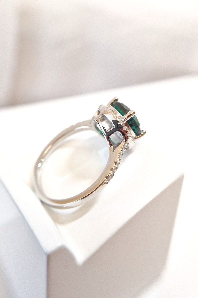 May include: A silver ring with a large, emerald green gemstone surrounded by smaller, clear gemstones. The ring is set on a white background.