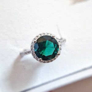 May include: A silver ring with a large, round, emerald green gemstone surrounded by a halo of smaller, clear gemstones.