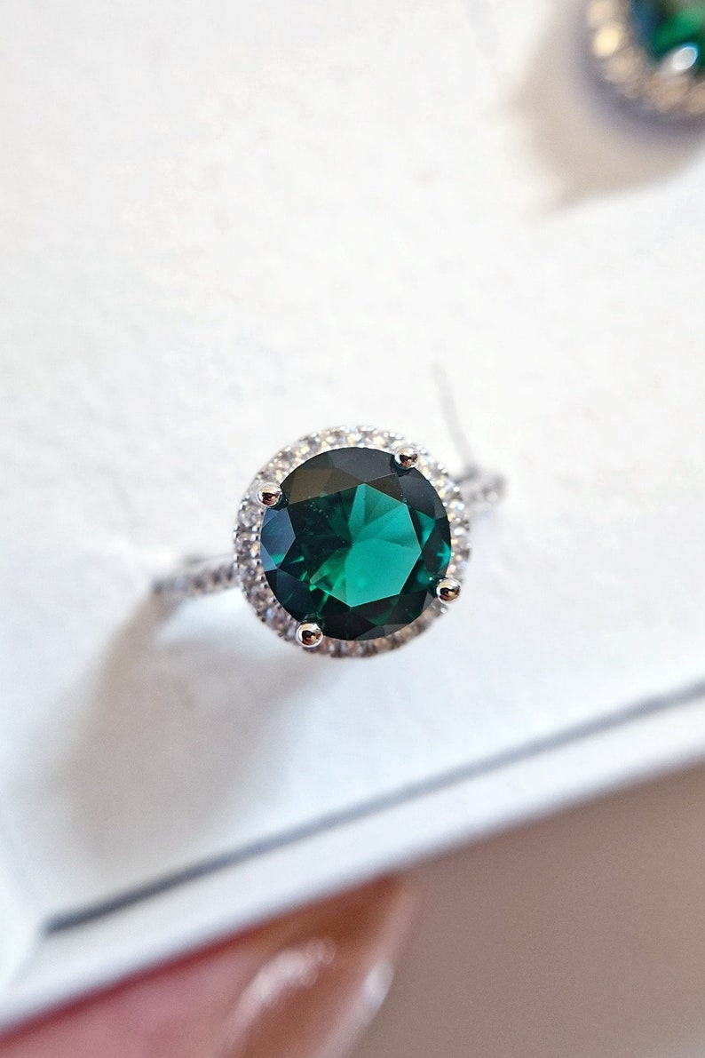 May include: A silver ring with a large, round, emerald green gemstone surrounded by a halo of smaller, clear gemstones.