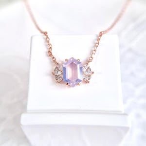 May include: A rose gold necklace featuring a hexagonal, light purple gemstone pendant. The pendant is flanked by two clusters of small, clear crystals. The necklace is displayed on a white surface.