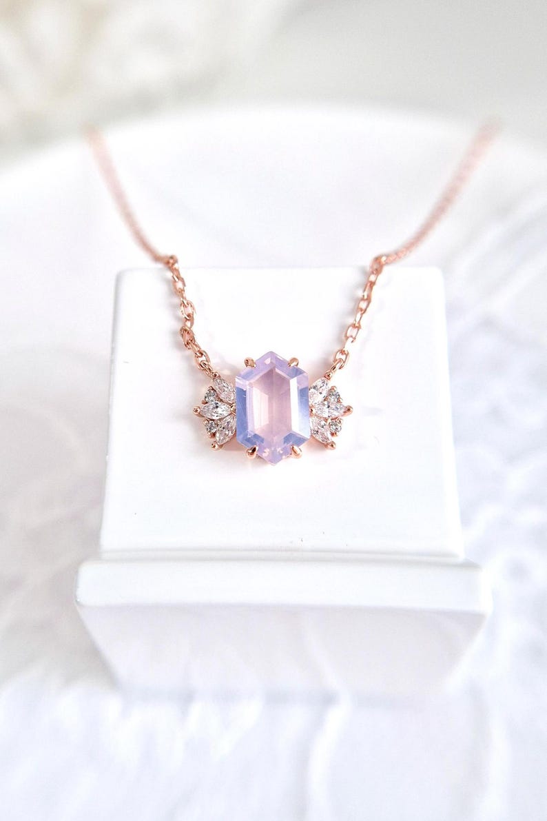 May include: A rose gold necklace featuring a hexagonal, light purple gemstone pendant. The pendant is flanked by two clusters of small, clear crystals. The necklace is displayed on a white surface.
