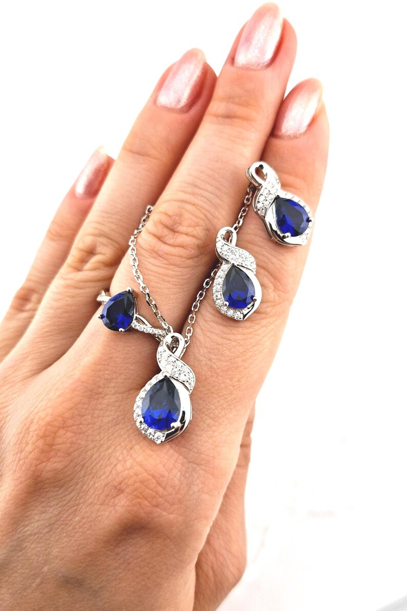 May include: A silver necklace, ring, and earrings set with a blue pear-shaped gemstone surrounded by smaller clear gemstones.