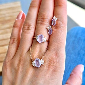 Peut inclure: Une main pr&eacute;sente un ensemble de bijoux comprenant une bague, un collier et des boucles d'oreilles. Les pi&egrave;ces dor&eacute;es pr&eacute;sentent des pierres pr&eacute;cieuses hexagonales violet clair avec des pierres d'accent blanches. La bague et le collier ont un motif floral.