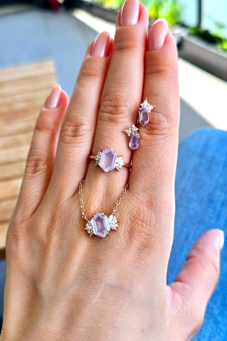 Peut inclure: Une main pr&eacute;sente un ensemble de bijoux comprenant une bague, un collier et des boucles d'oreilles. Les pi&egrave;ces dor&eacute;es pr&eacute;sentent des pierres pr&eacute;cieuses hexagonales violet clair avec des pierres d'accent blanches. La bague et le collier ont un motif floral.